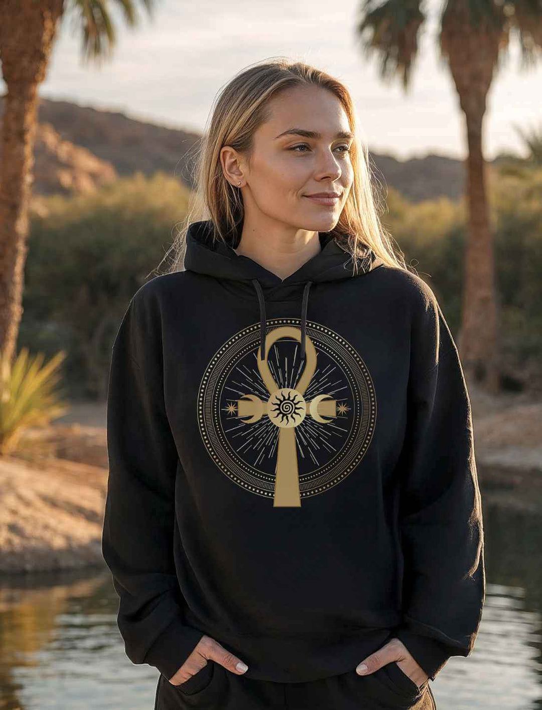 Frau mit blondem Haar trägt im Sonnenuntergang einen schwarzen Hoodie mit goldenem Ankh-Symbol und Dreifachmond-Design. Palmen und Wüstenszenerie im Hintergrund.
