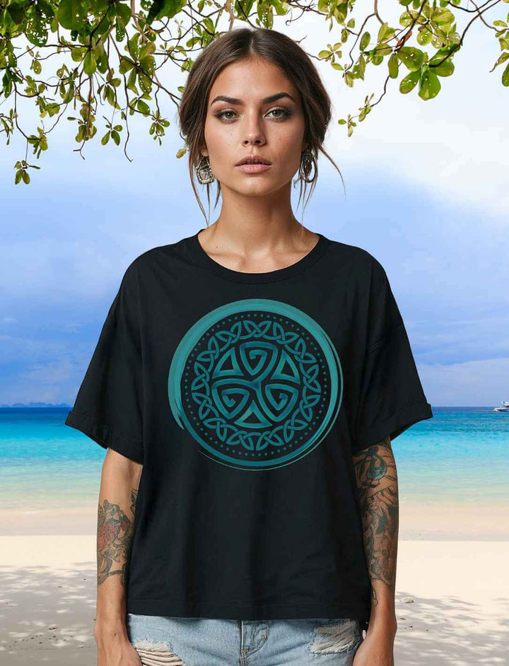 Locker geschnittenes Damen Bio-Shirt in Schwarz mit türkisfarbener Triskele auf der Brust – mystisch & kraftvoll am Strand inszeniert.