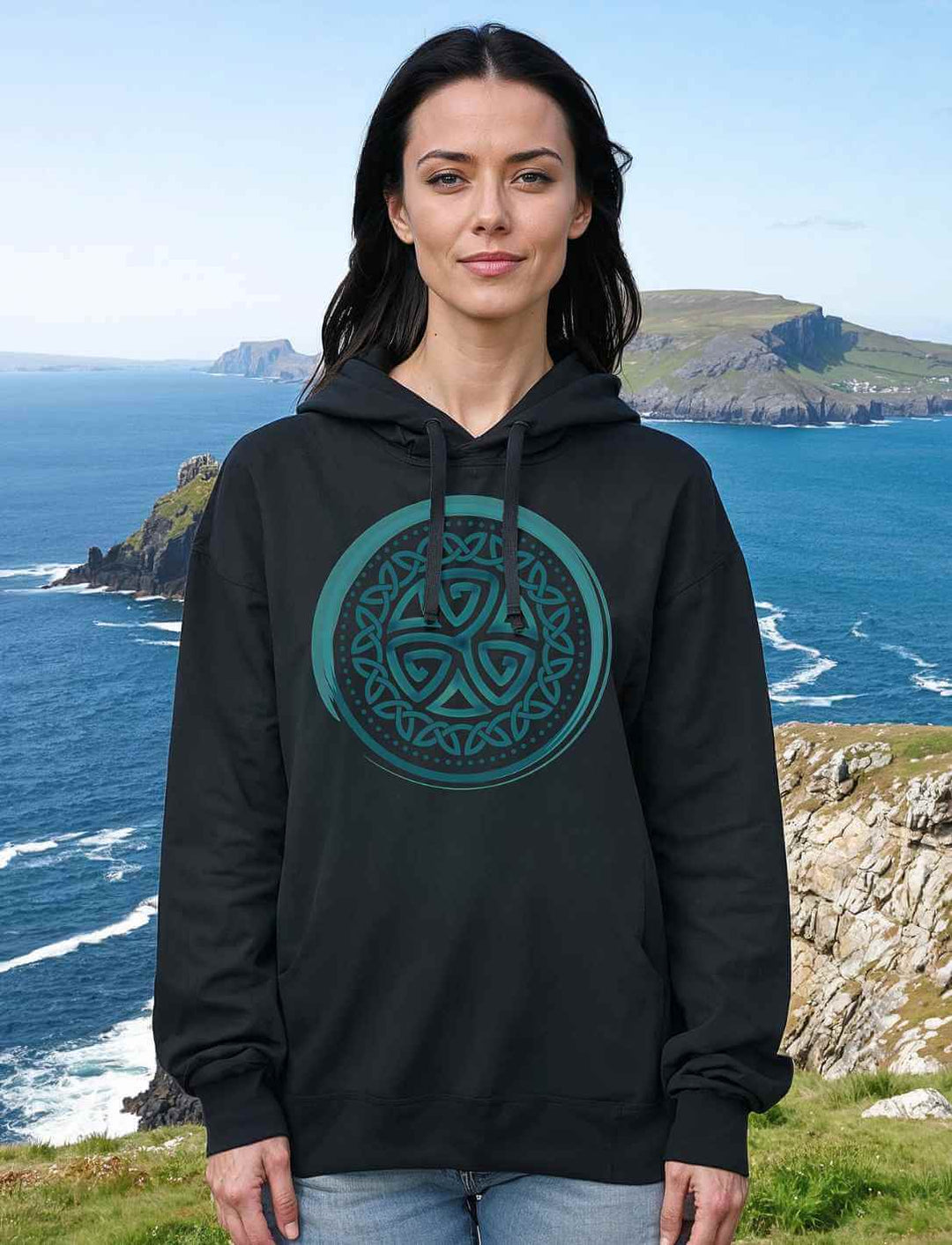 Frau trägt den Aqua Triskele Hoodie in Schwarz, mit keltischem Symbol, vor einer rauen Küstenlandschaft