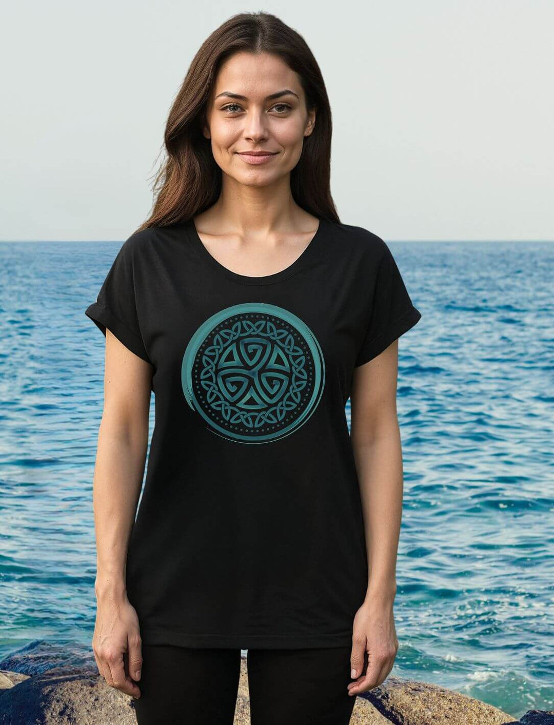 Frau am Meer trägt schwarzes Damen Longfit Shirt mit türkisfarbener keltischer Triskele im Kreisdesign.