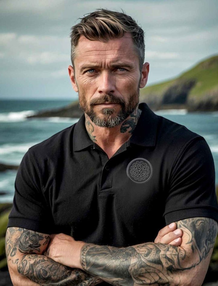Mann mit tätowierten Armen trägt ein schwarzes Unisex Organic Poloshirt „Aqua Triskele“ mit gesticktem Emblem, stehend vor einer Küstenlandschaft.