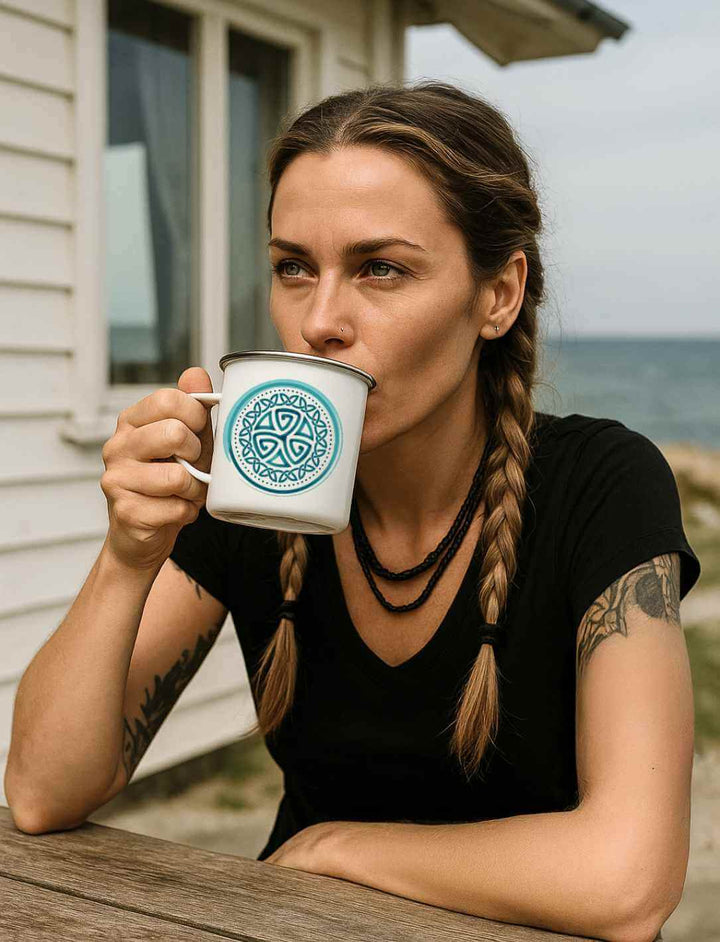 Junge Frau mit Tattoos trinkt aus einer weißen Emaille-Tasse „Aqua Triskele“ mit silbernem Rand am Tisch vor einem Haus am Meer.
