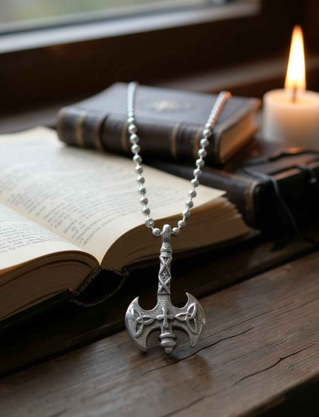 Edelstahl-Anhänger „Axt des Sonnenkreuzes“ auf Kette, liegend auf aufgeschlagenem Buch mit Kerzenschein an Edelstahlkette silberfarben.