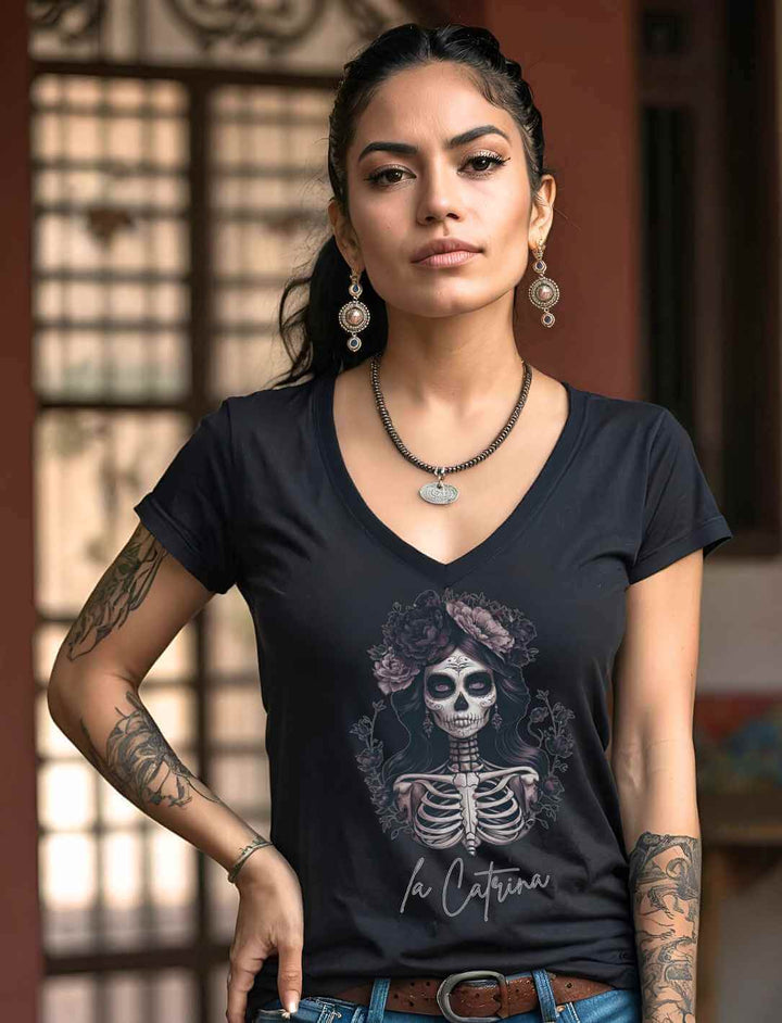 Junge Frau trägt das Catrina Deluxe Damen V-Neck Shirt in Schwarz mit La Catrina Design, fotografiert in einem stilvollen mexikanischen Innenhof.