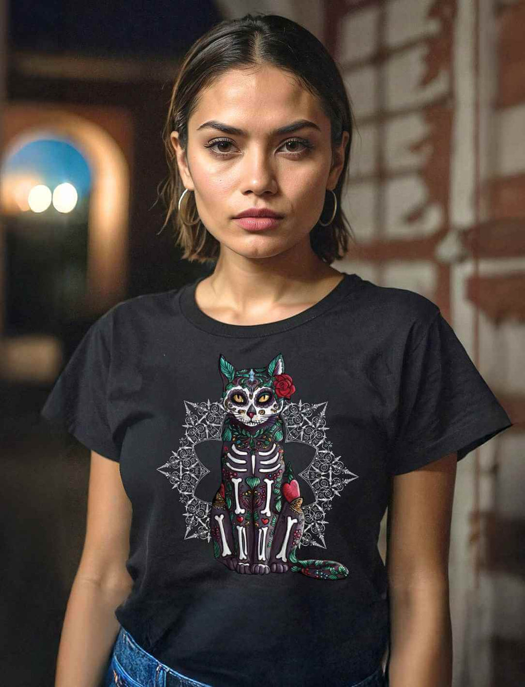 Junge Frau trägt Catrina Felina Damen T-Shirt in Schwarz – Mystische Katzenkunst im Stil des Día de los Muertos, elegante Produktabbildung vor einem abendlichen Hintergrund.
