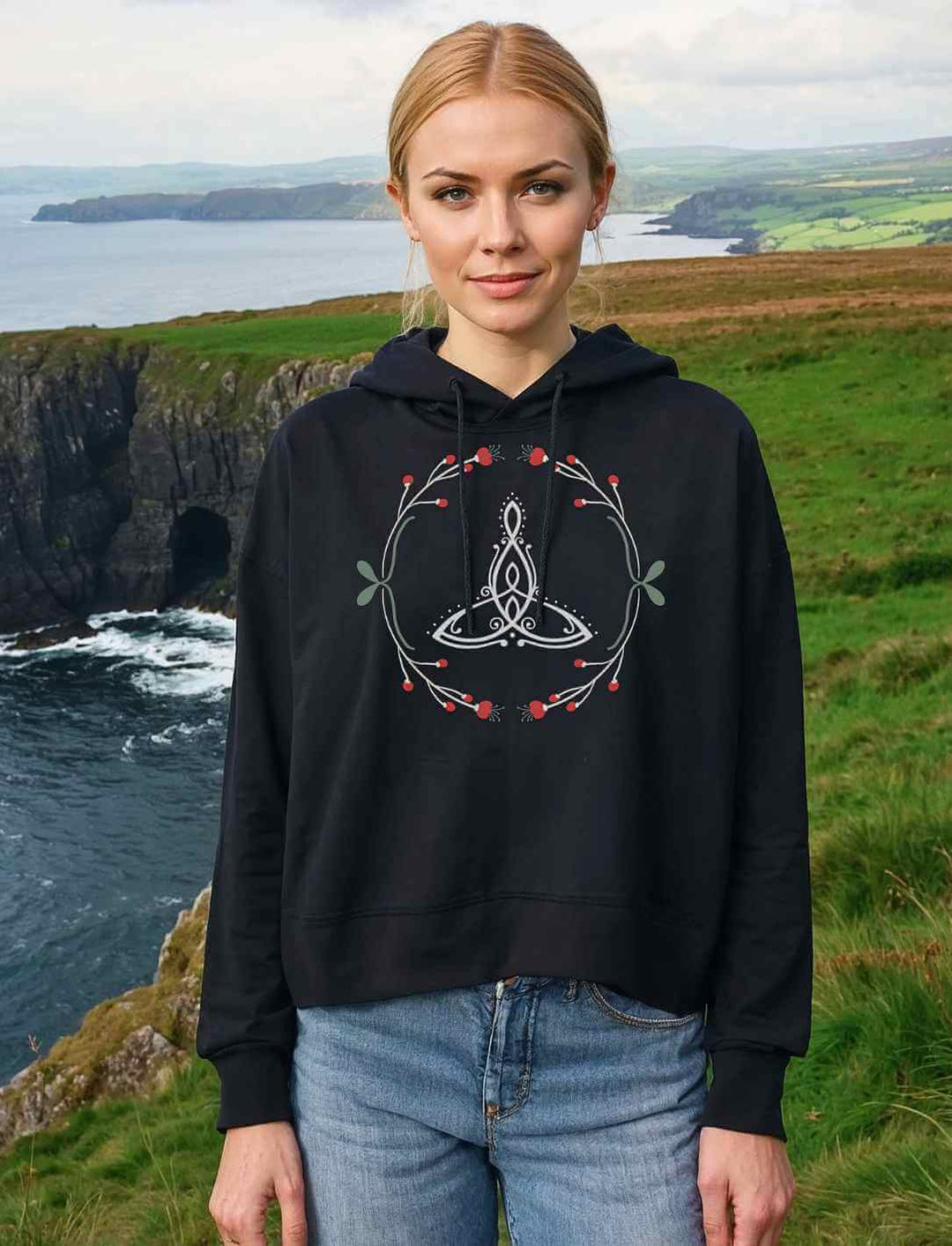 Eine Frau steht auf einer grĂŒnen Klippe mit Blick auf das raue Meer und trĂ€gt das Celtic Mother Damen Boxcut Hoodie in Schwarz. Das filigrane Triquetra-Motiv mit zarten Naturverzierungen verleiht dem Look eine mystische Aura.