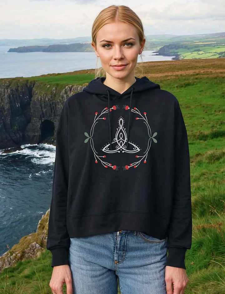 Eine Frau steht auf einer grĂŒnen Klippe mit Blick auf das raue Meer und trĂ€gt das Celtic Mother Damen Boxcut Hoodie in Schwarz. Das filigrane Triquetra-Motiv mit zarten Naturverzierungen verleiht dem Look eine mystische Aura.