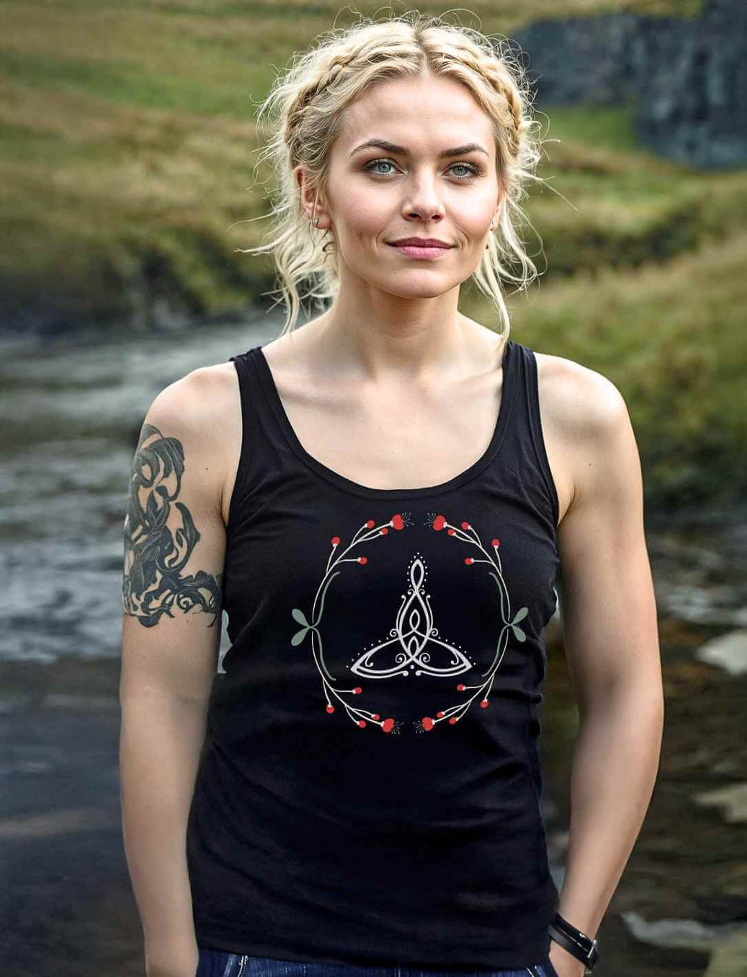 Frau mit blonden Zöpfen und Tattoo trĂ€gt das âCeltic Motherâ Tank-Top in Schwarz vor einem Bach in der Natur.