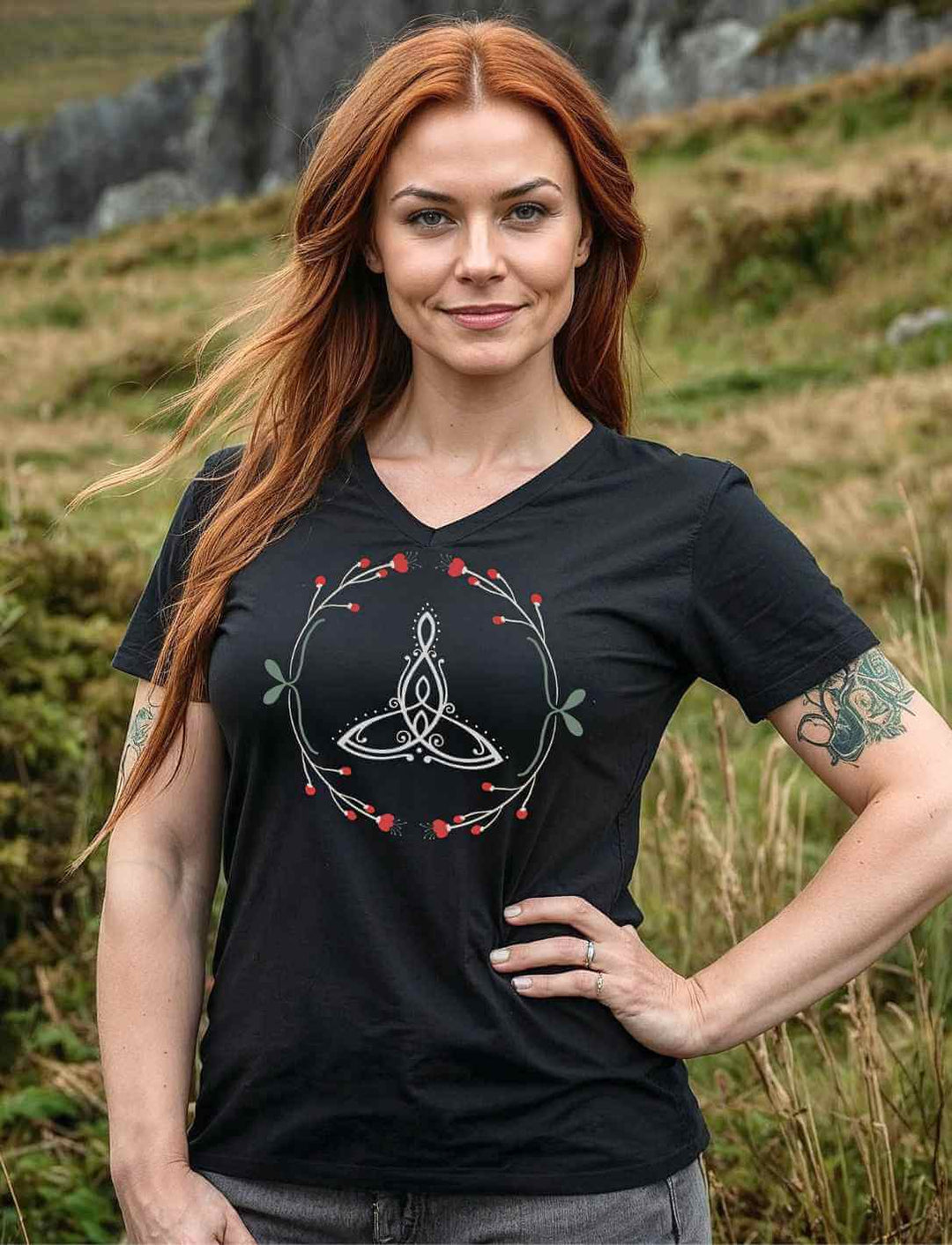 Keltische Frau mit roten Haaren trĂ€gt das âCeltic Motherâ V-Neck-Shirt in Schwarz vor einer irischen Landschaft.