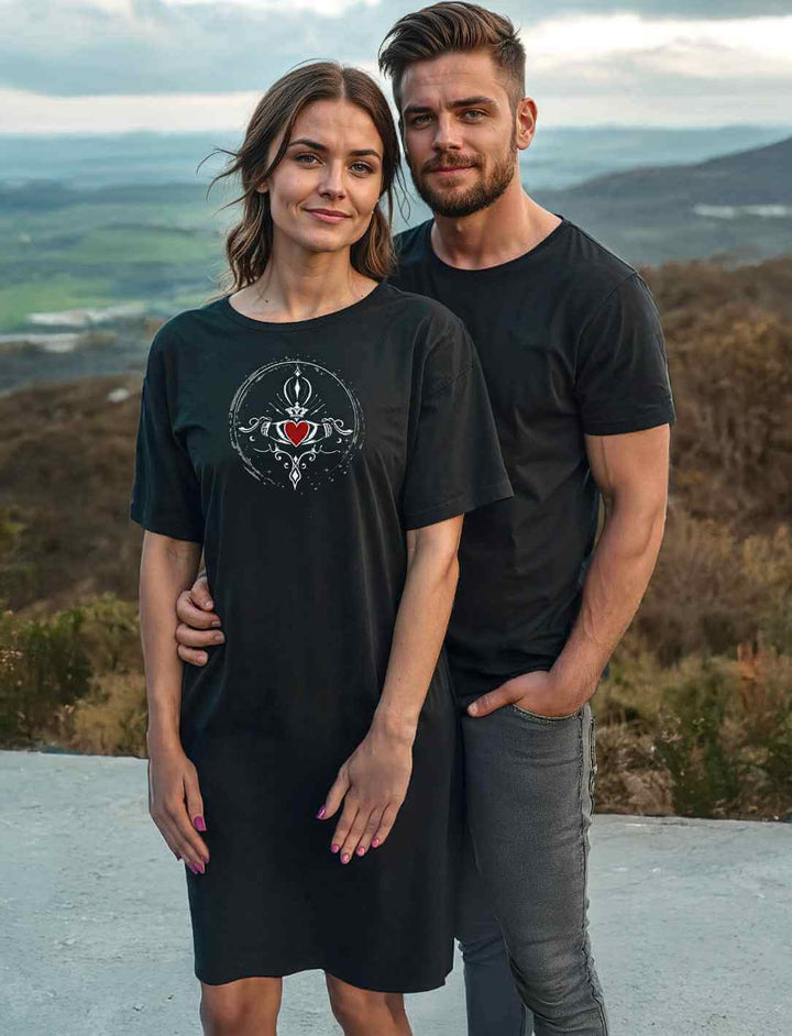 Model trägt Claddagh Damen Kleid in der Natur – Ein Paar, das das Claddagh T-Shirt Kleid präsentiert. Die Frau trägt das schwarze Kleid mit Claddagh-Motiv, umgeben von einer idyllischen Landschaft.