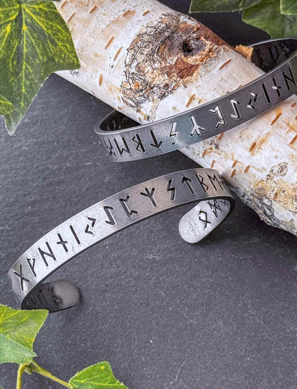 Zwei Runental Edelstahl Armreife mit nordischer Runengravur auf Birkenholz, vollständiges Futhark mit Efeudekoration.