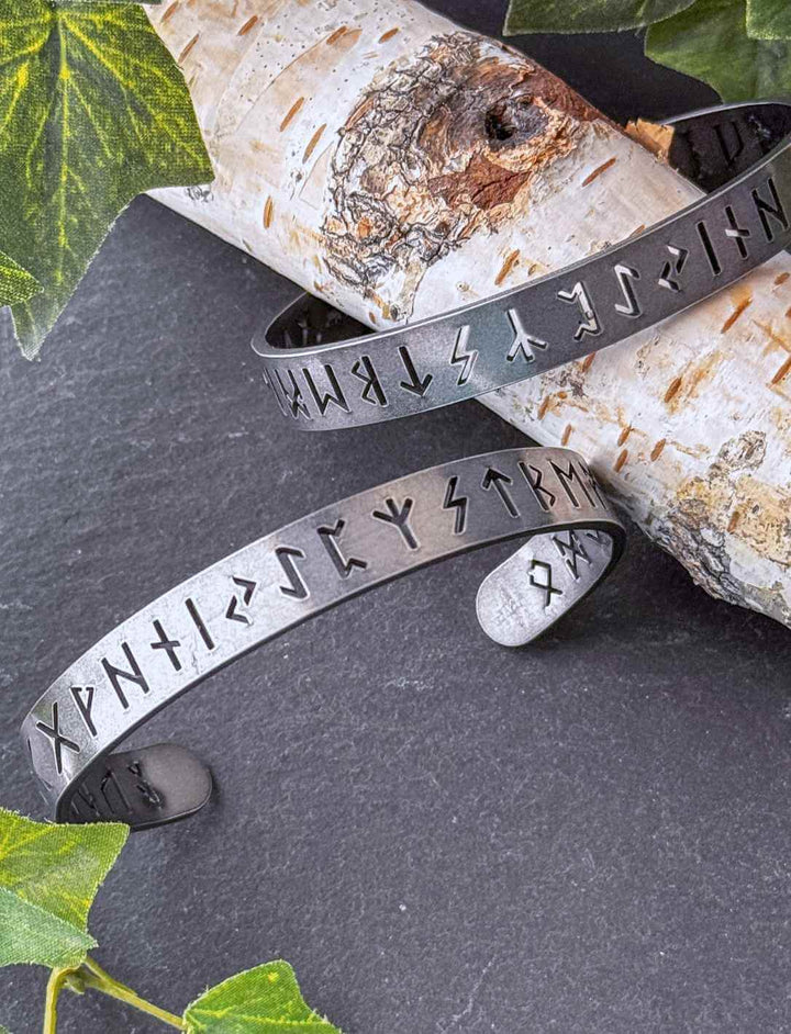 Zwei Runental Edelstahl Armreife mit nordischer Runengravur auf Birkenholz, vollständiges Futhark mit Efeudekoration.