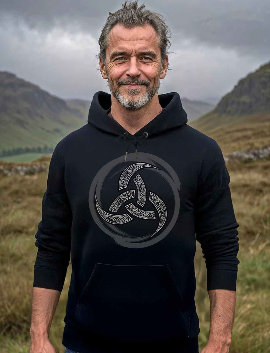 Ein Mann mittleren Alters mit zurückgebundenen grauen Haaren und Bart trägt einen schwarzen Hoodie mit silbernem Triquetra-Symbol. Er steht lächelnd in einer nebligen, grünen Hügellandschaft.