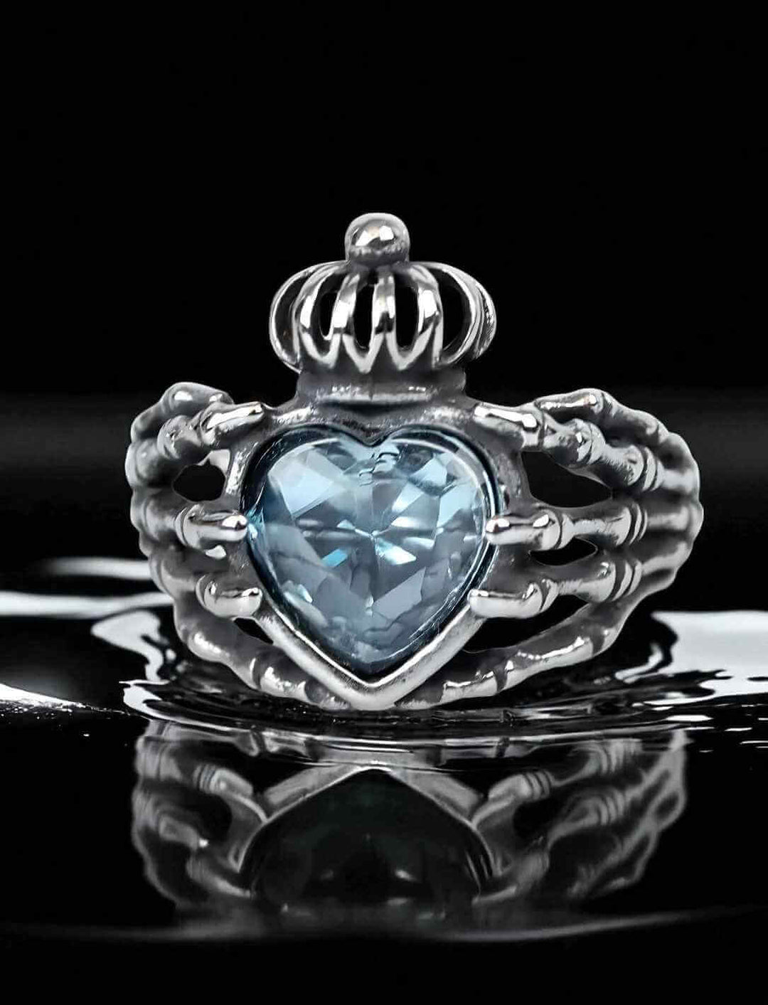 Frontansicht des Claddagh-Rings "Ewiges Versprechen" mit Skelett-Händen und eisblauem Herzstein auf schwarzem Spiegelgrund.