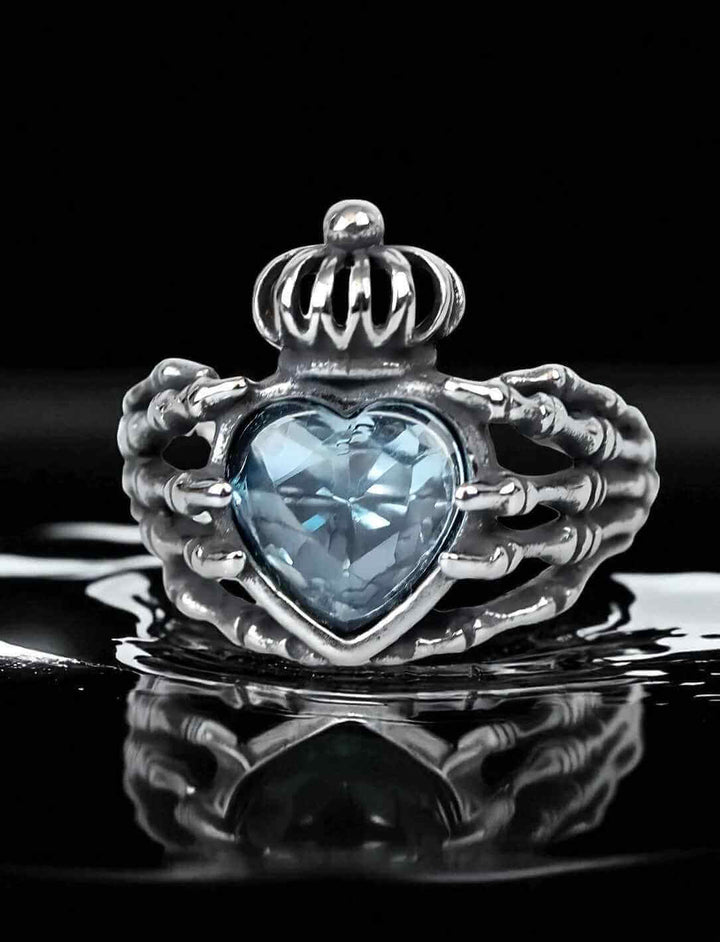 Frontansicht des Claddagh-Rings "Ewiges Versprechen" mit Skelett-Händen und eisblauem Herzstein auf schwarzem Spiegelgrund.