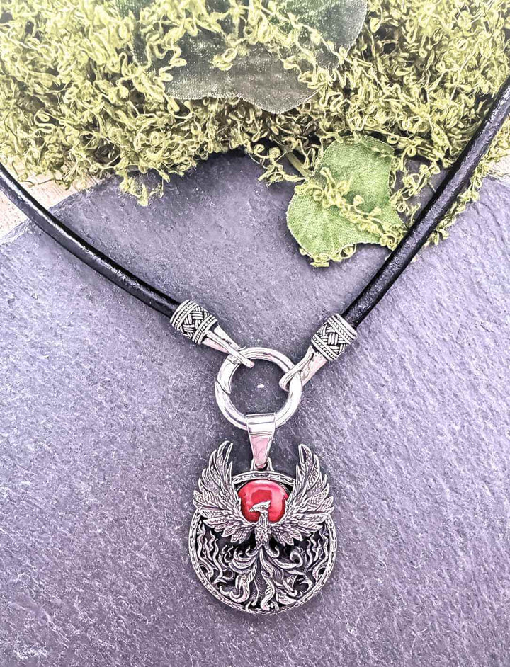 Phönix-Anhänger an Lederband auf grauer Stein Unterlage – Edelstahl mit rotem Glasstein kombiniert, mit Lederband des Hüters.