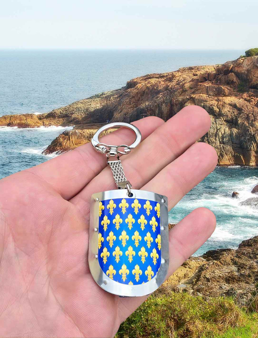 Der 'Fleur de Lis Schild' Schlüsselanhänger, fest in einer Hand vor malerischem Küstenpanorama mit dem Meer im Hintergrund - ein Symbol der Freiheit und Geschichte von Runental.de.