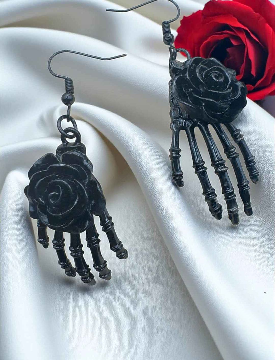 La Rosa Oscura – Skelett-Ohrringe mit schwarzer Rose, dekorativ präsentiert auf weißem Satin neben einer roten Rose – Runental Schmuckstück im Día de los Muertos-Stil