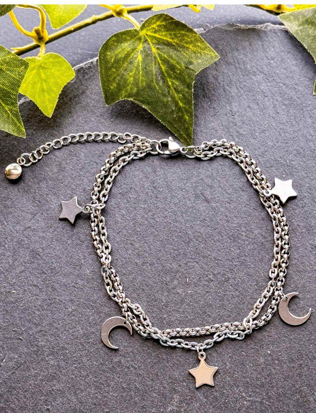 Lunaria Fußkettchen aus Edelstahl mit Monden und Sternen, fotografiert auf dunkler Steinplatte unter grünen Efeublättern – Runental Schmuckstück
