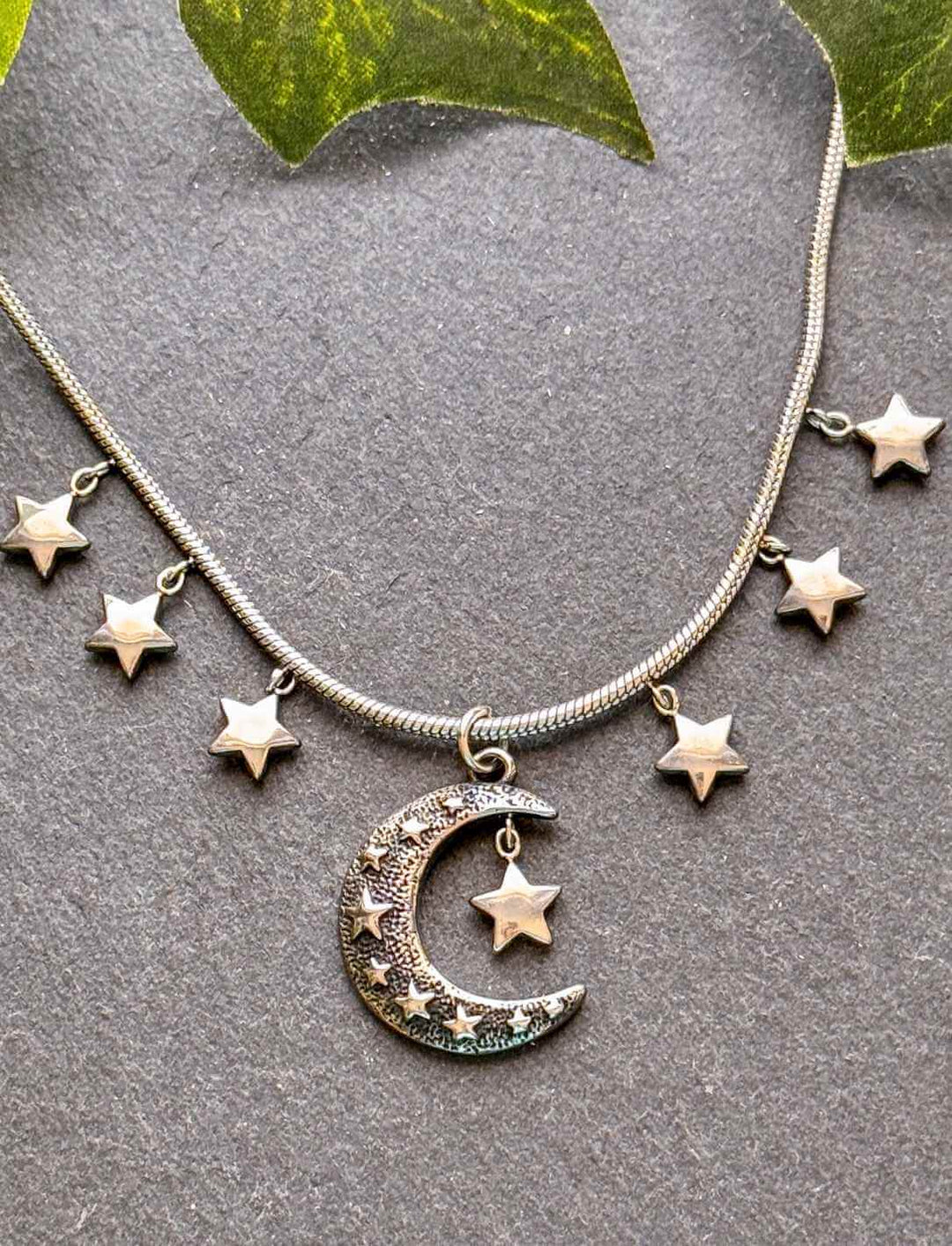 Lunaria Edelstahlkette mit Mond & Sternen – filigraner Fantasy-Schmuck auf grauem Untergrund mit Blättern