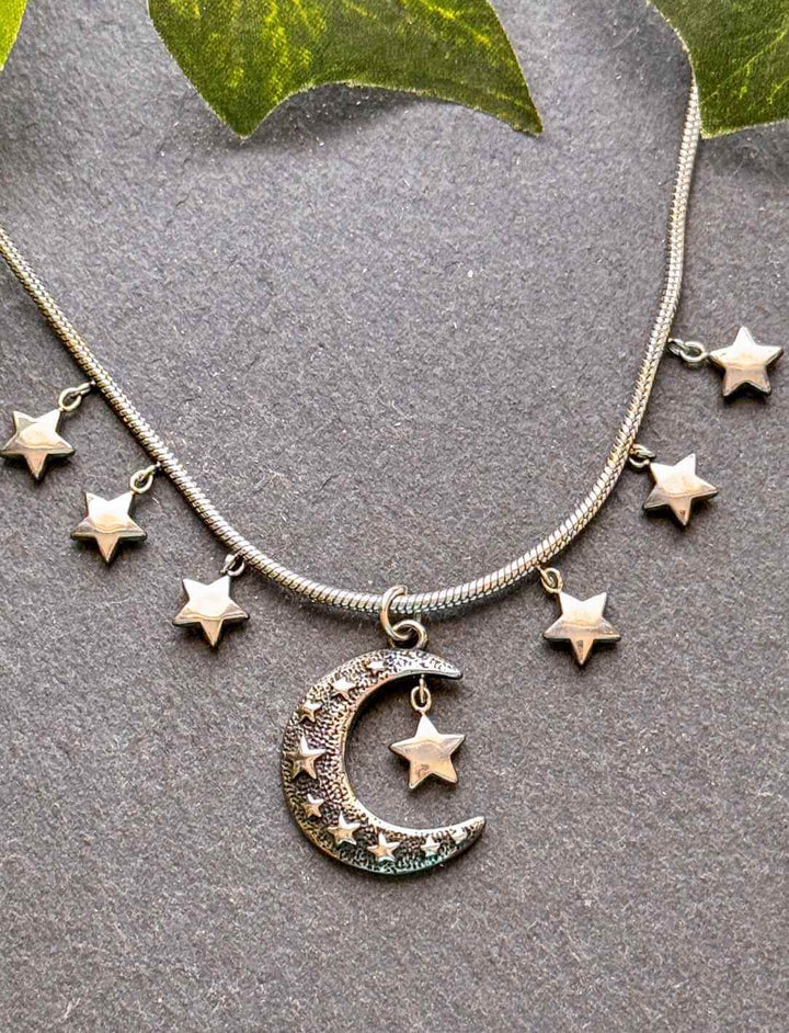 Lunaria Edelstahlkette mit Mond & Sternen – filigraner Fantasy-Schmuck auf grauem Untergrund mit Blättern