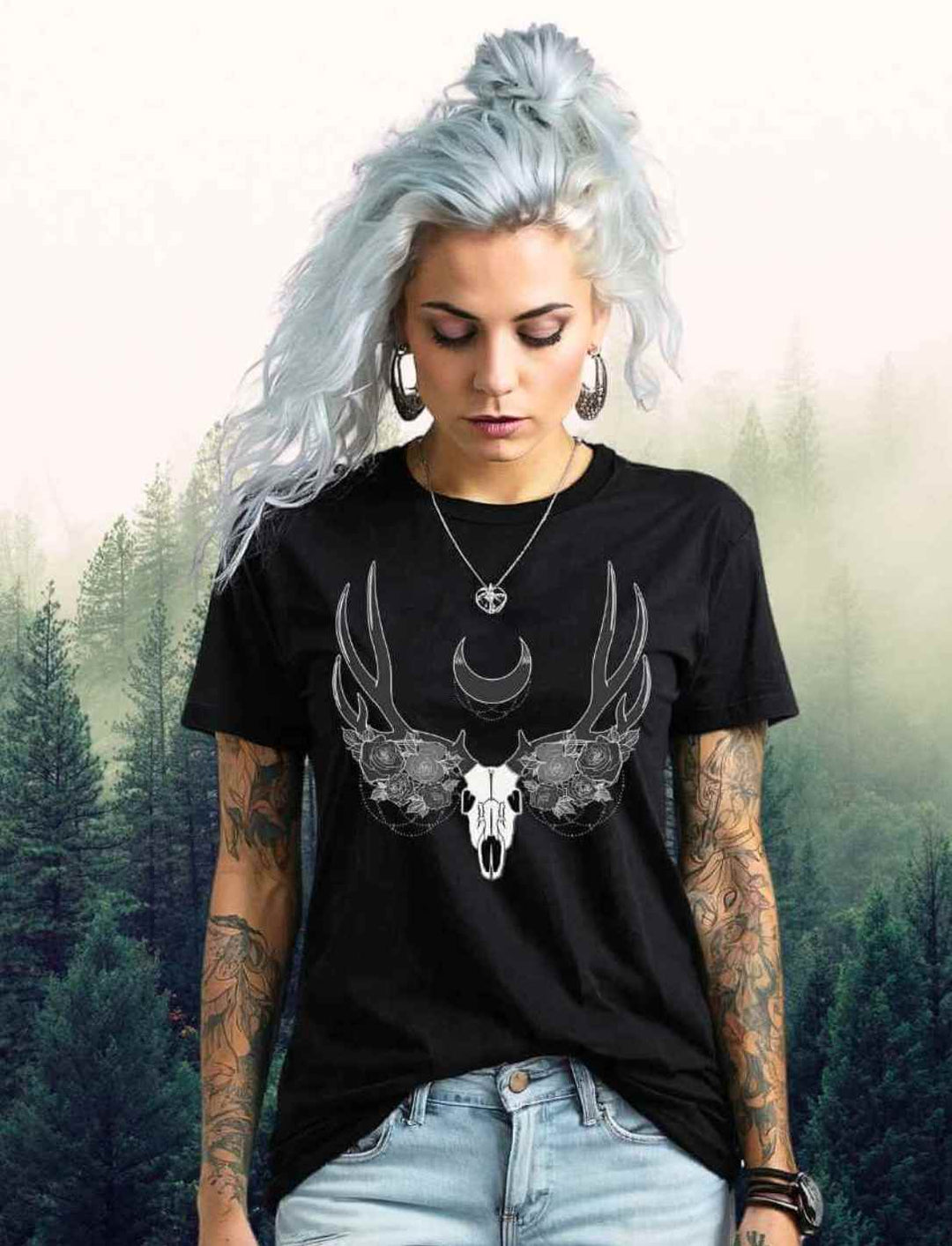 Ein weibliches Model mit eisblauem Haar trägt ein schwarzes Lunas Mondgeweih T-Shirt vor einem mystischen Waldhintergrund, die Ästhetik ergänzt durch ihre sichtbaren Arm-Tattoos.