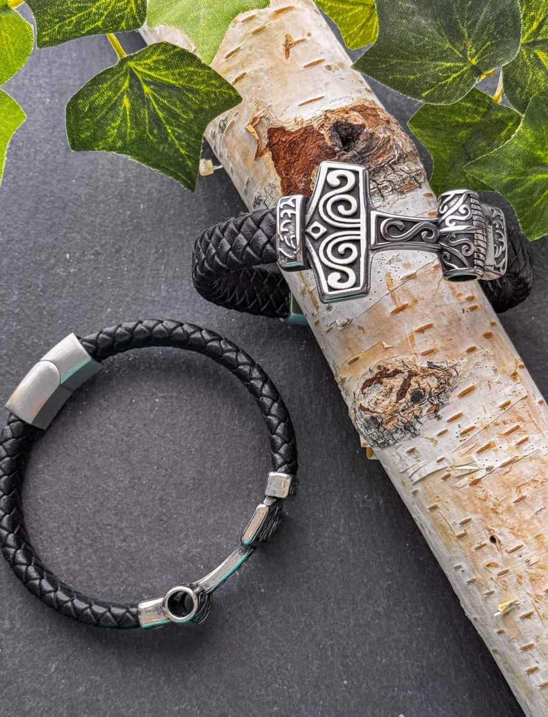 Lederarmband „Mjölnirs Griff“ mit Edelstahlverschluss und Thorshammer-Motiv, arrangiert auf Birkenholz und grünem Efeu
