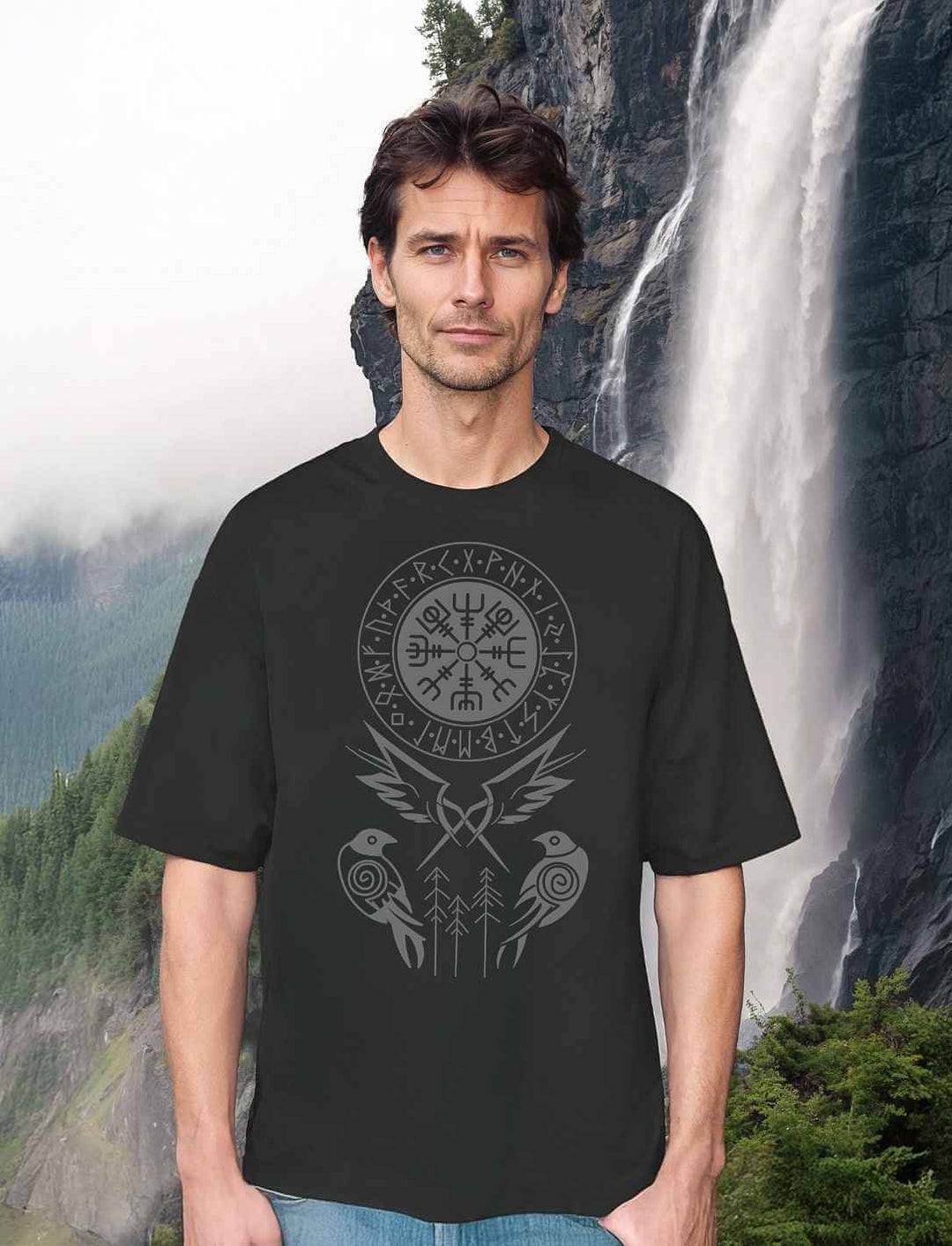 Model trägt Oversized T-Shirt „Nordwind – Die Raben Odins“ in Schwarz mit Vegvisir, Runenkreis und zwei stilisierten Raben vor Wasserfallkulisse.