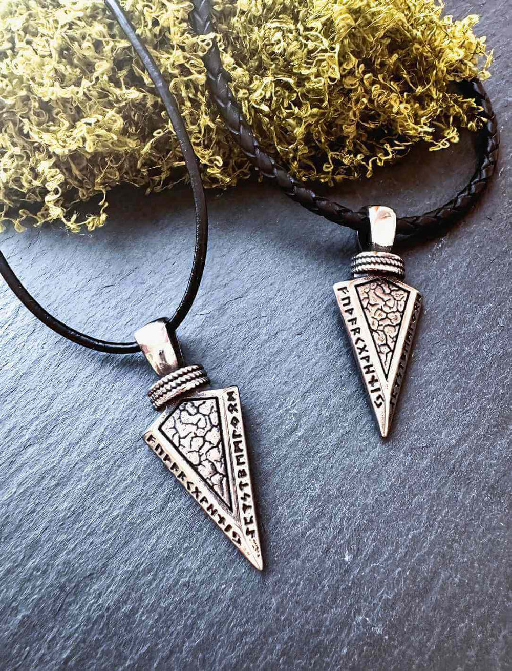 Zwei Edelstahl-Amulette in Pfeilform mit Runeninschrift, auf schwarzem Hintergrund mit Moos und Lederband
