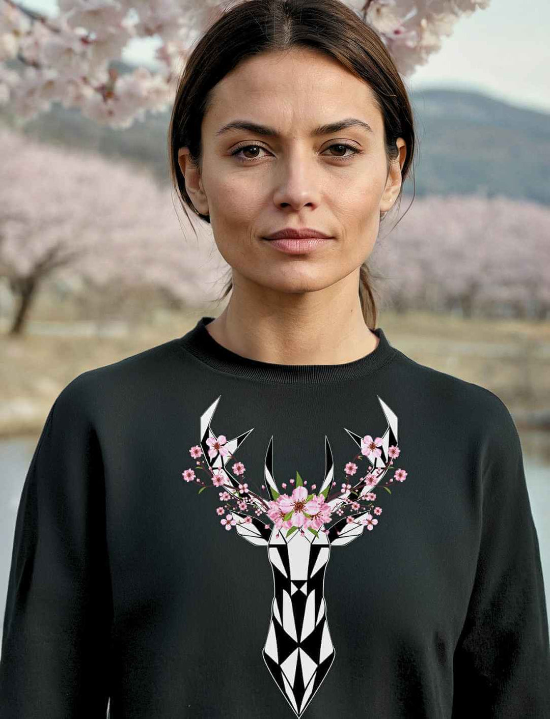 Eine Frau trägt den schwarzen Sakura Deer Organic Unisex Sweater vor einem Hintergrund aus blühenden Kirschblüten, der die japanische Eleganz und Naturverbundenheit widerspiegelt.