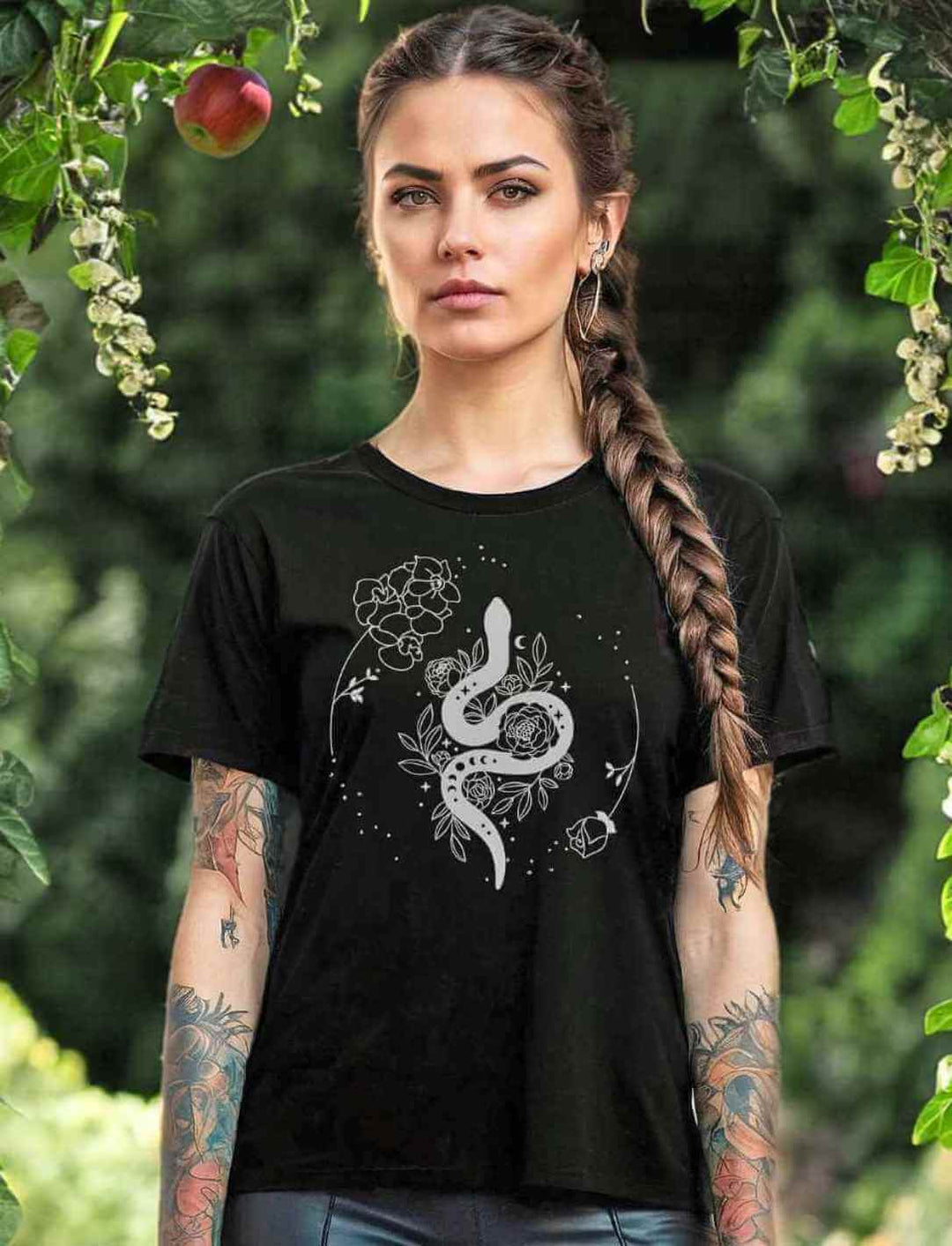 Junge Frau mit Zopffrisur trägt das 'Snake of Wisdom' T-Shirt in Schwarz, das mit einem weißen Schlangen- und Blumenmuster bedruckt ist, vor einem üppigen grünen Gartenhintergrund, der die Verbindung zur Natur unterstreicht