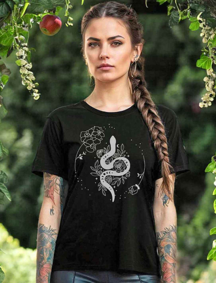 Junge Frau mit Zopffrisur trägt das 'Snake of Wisdom' T-Shirt in Schwarz, das mit einem weißen Schlangen- und Blumenmuster bedruckt ist, vor einem üppigen grünen Gartenhintergrund, der die Verbindung zur Natur unterstreicht