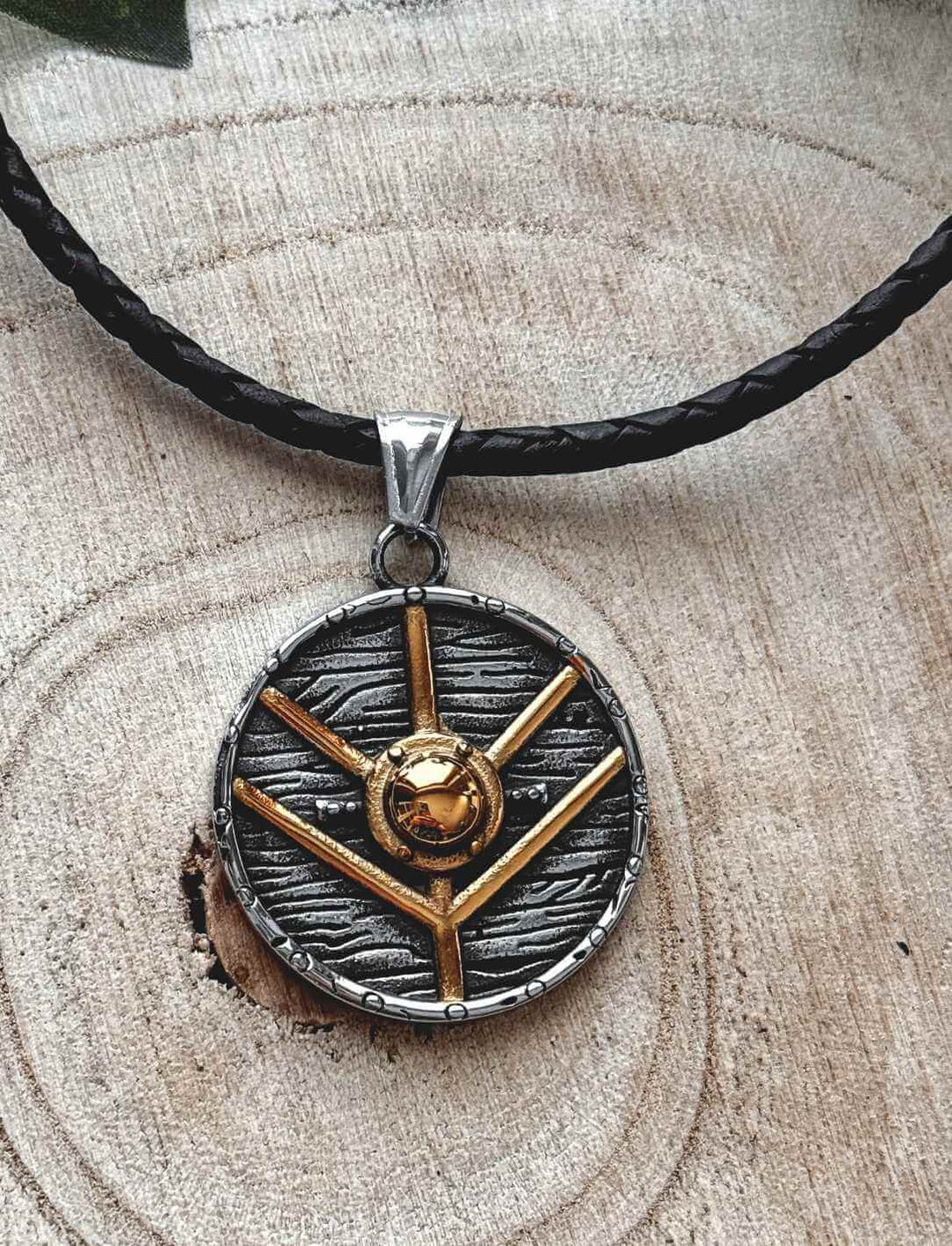 Runenverzierter Titan-Anhänger mit goldfarbener Schildform – Symbol aus der nordischen Mythologie, auf dekorativem Holzhintergrund mit geflochtener Lederkette.