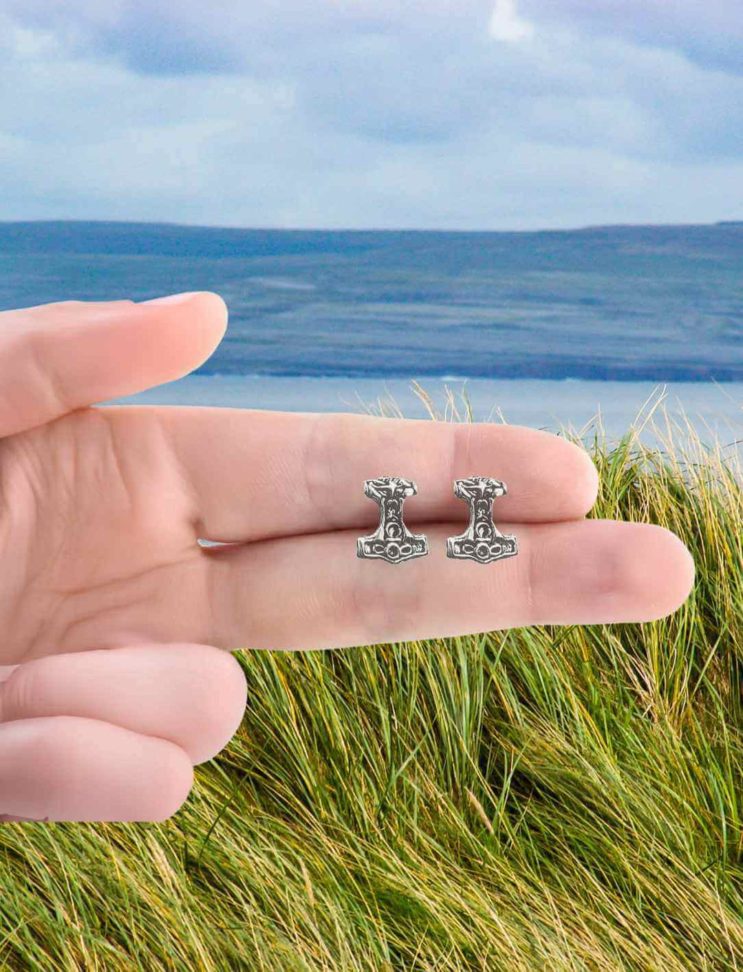 Nahaufnahme von Thor’s Hammer Ohrsteckern aus 925er Silber, gehalten vor einer malerischen nordischen Landschaft. Symbol für Stärke und Schutz in der nordischen Mythologie.