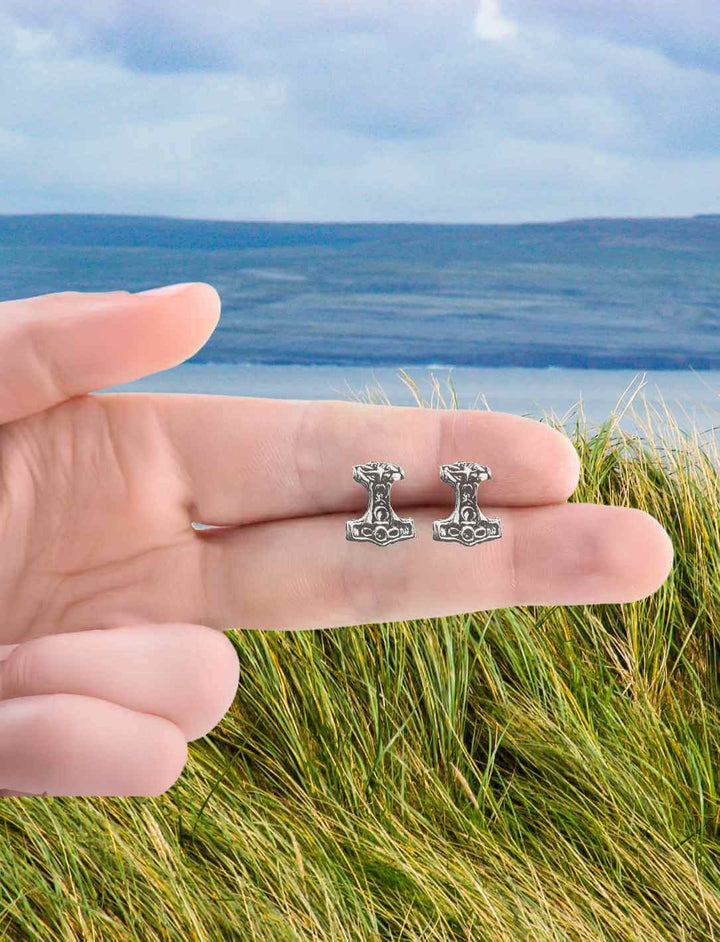 Nahaufnahme von Thor’s Hammer Ohrsteckern aus 925er Silber, gehalten vor einer malerischen nordischen Landschaft. Symbol für Stärke und Schutz in der nordischen Mythologie.