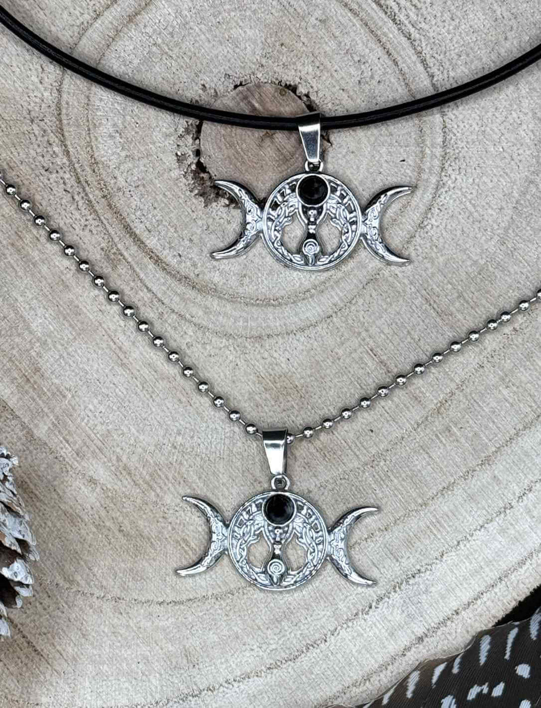 Triple Moon – Wicca Amulett mit Onyx, einmal an Edelstahl-Kugelkette und einmal an schwarzer Lederkette auf Holz präsentiert.