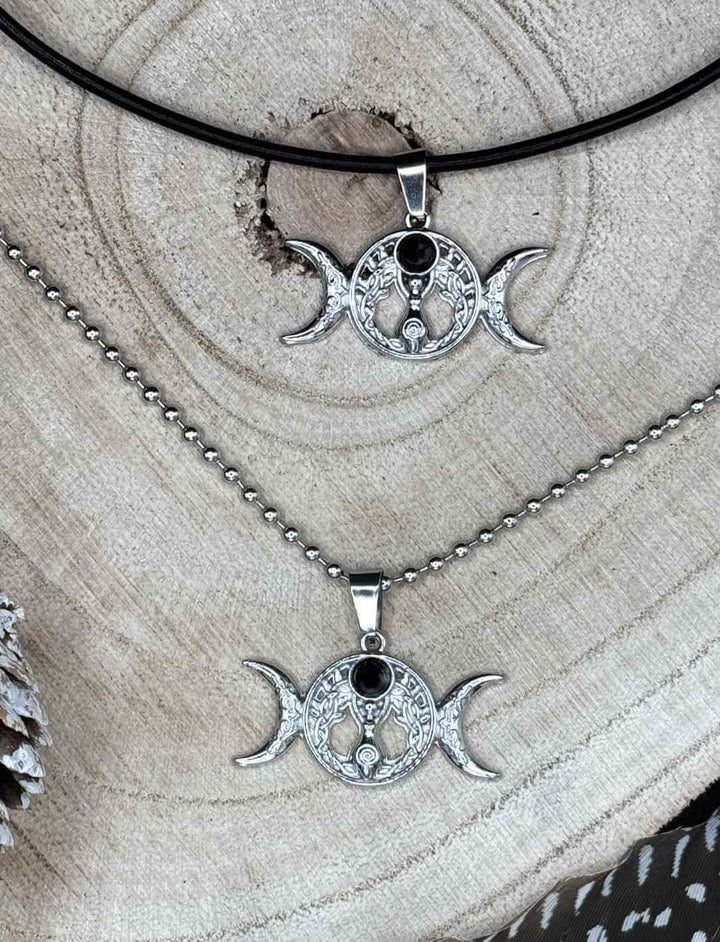 Triple Moon – Wicca Amulett mit Onyx, einmal an Edelstahl-Kugelkette und einmal an schwarzer Lederkette auf Holz präsentiert.