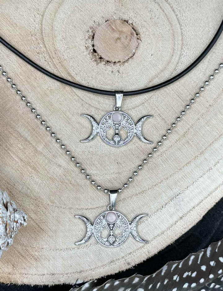 Triple Moon – Wicca Amulett mit Rosenquarz, einmal an Edelstahl-Kugelkette und einmal an schwarzer Lederkette auf Holz präsentiert.