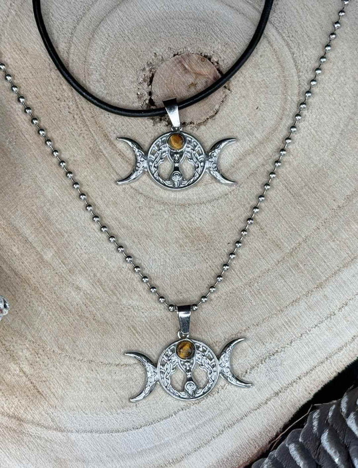 Triple Moon – Wicca Amulett mit Tigerauge, einmal an Edelstahl-Kugelkette und einmal an schwarzer Lederkette auf Holz präsentiert.