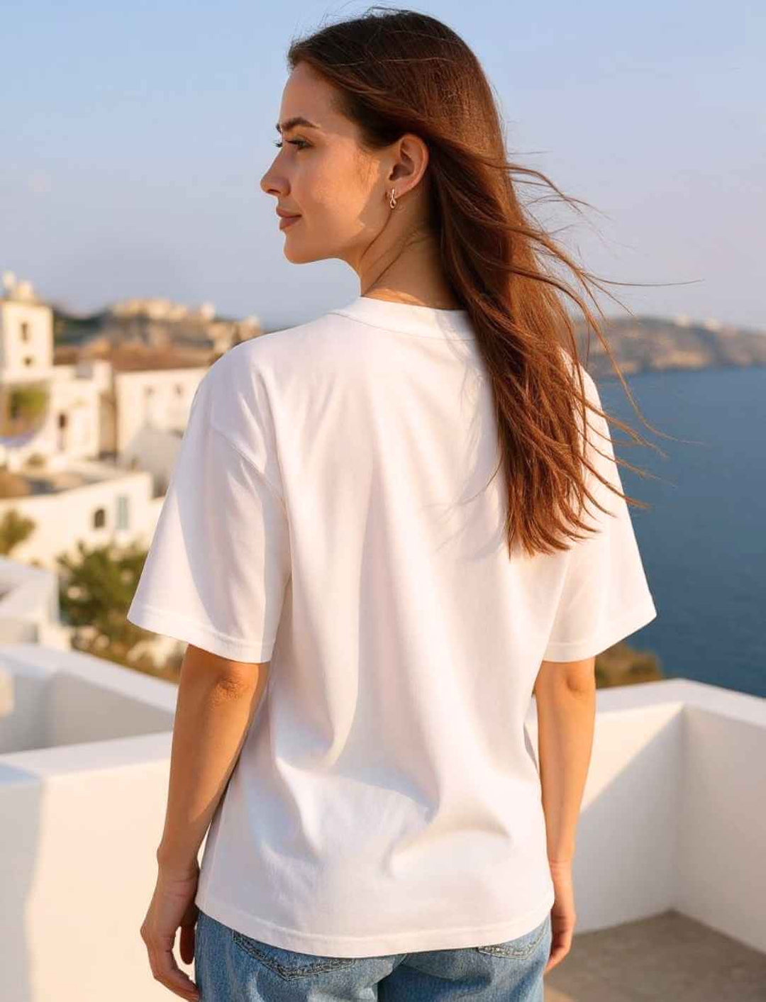 Model von hinten mit weißem Oversized Bio-T-Shirt – lockere Passform mit überschnittenen Ärmeln, fotografiert bei Sonnenuntergang auf einer mediterranen Dachterrasse.