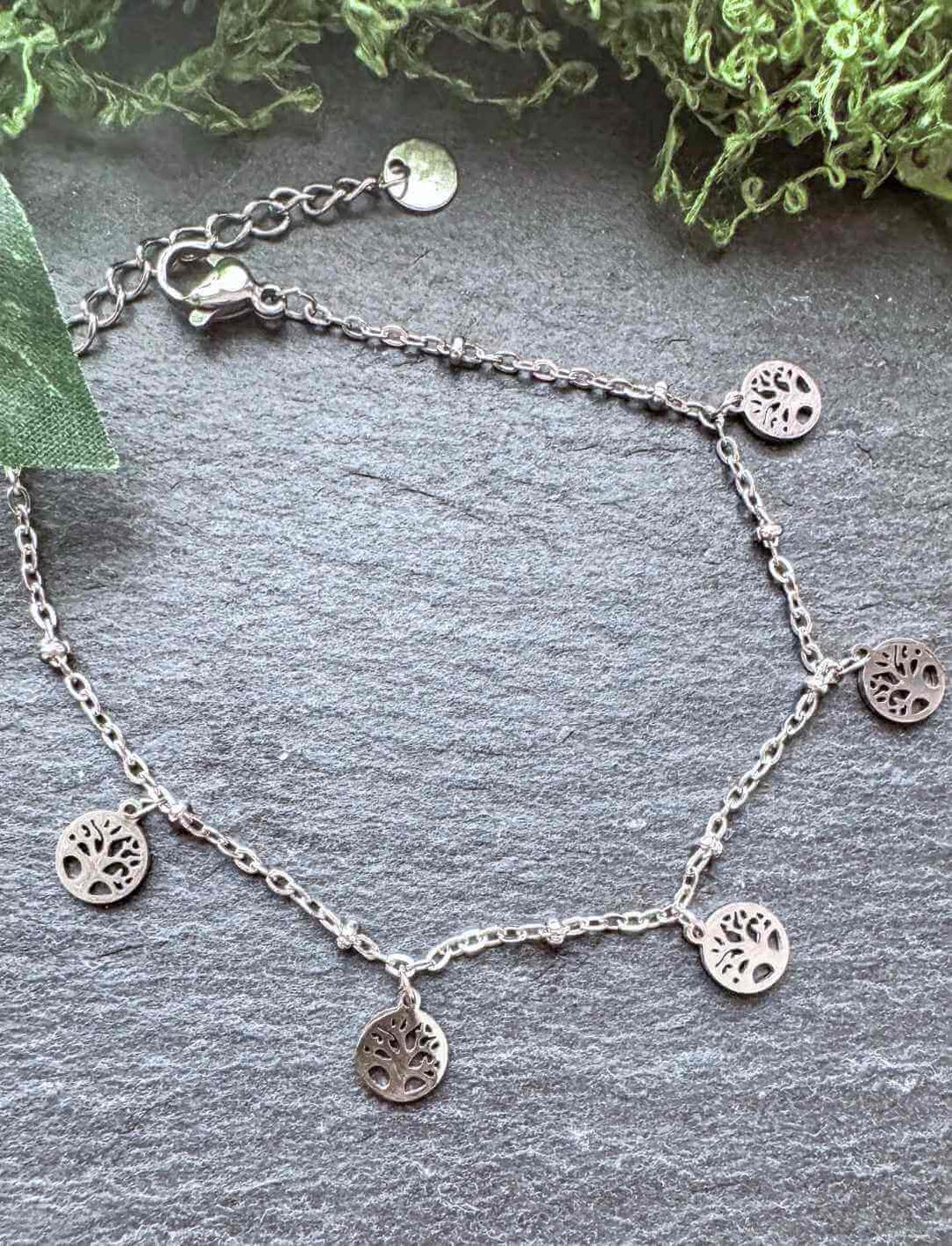 Filigranes Damenarmband aus Edelstahl mit fünf baumelnden Lebensbaum-Anhängern, inspiriert von der keltischen Göttin Danu – fotografiert auf dunklem Schiefer mit moosigem Dekor.