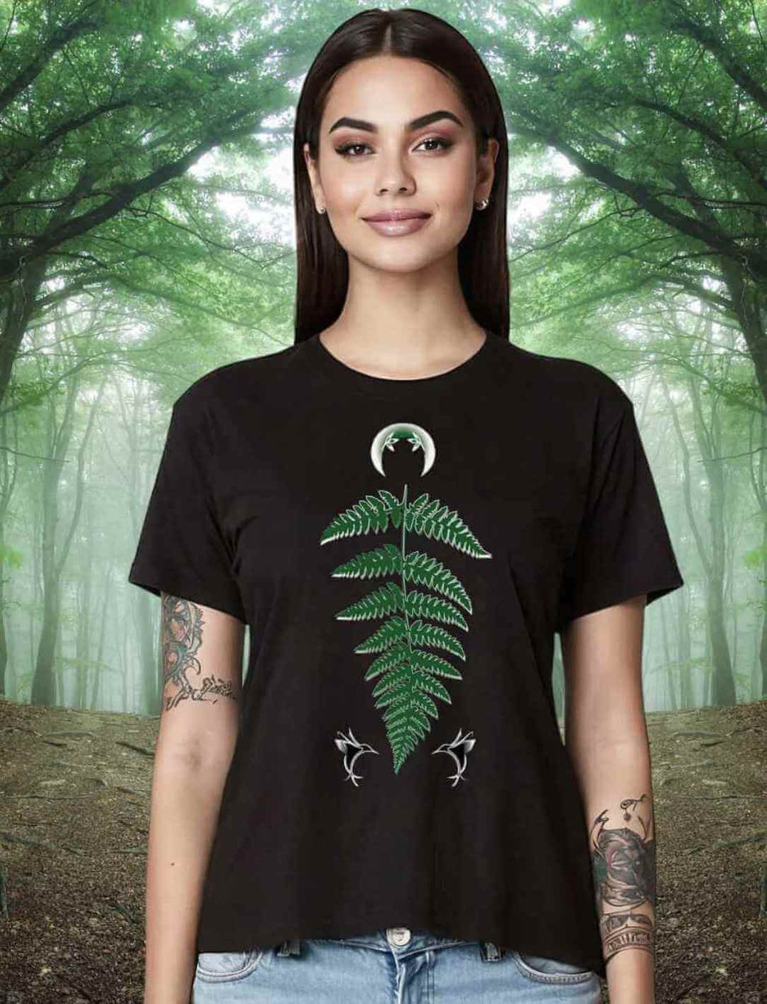 Frau mit langen dunklen Haaren trägt ein schwarzes Damen-T-Shirt 'Zauberhafte Naturwelt' mit grünem Farnblatt- und weißen Libellen-Motiv, vor einem Waldhintergrund posierend.