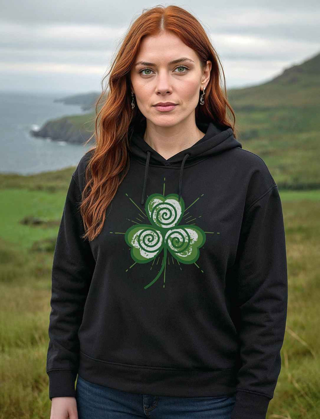 Rothaarige Frau trägt schwarzen Organic Boxcut Hoodie mit kleeblattförmiger Triskele vor irischer  Naturkulisse