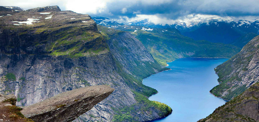 Spektakuläre Aussicht auf Felsformation in Norwegen, hoch über einem tiefblauen Fjord, umgeben von steilen Bergen und teilweise schneebedeckten Gipfeln.