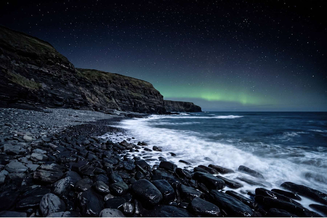 Nächtliche Nordseeküste mit schwarzen Schieferklippen und nassen dunklen Steinen unter Sternenhimmel mit Polarlicht – die Küste, an der Wikinger Gagat fanden