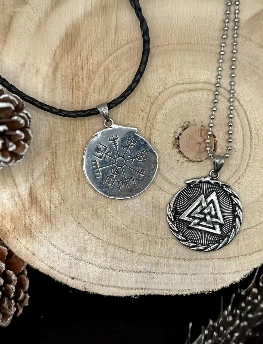 Amulett der nordischen Pfade – doppelseitiger Edelstahlanhänger mit Valknut und Vegvisir, kombiniert mit einer geflochtenen Lederkette und einer Kugelkette auf einer Holzscheibe, umgeben von Kiefernzapfen.