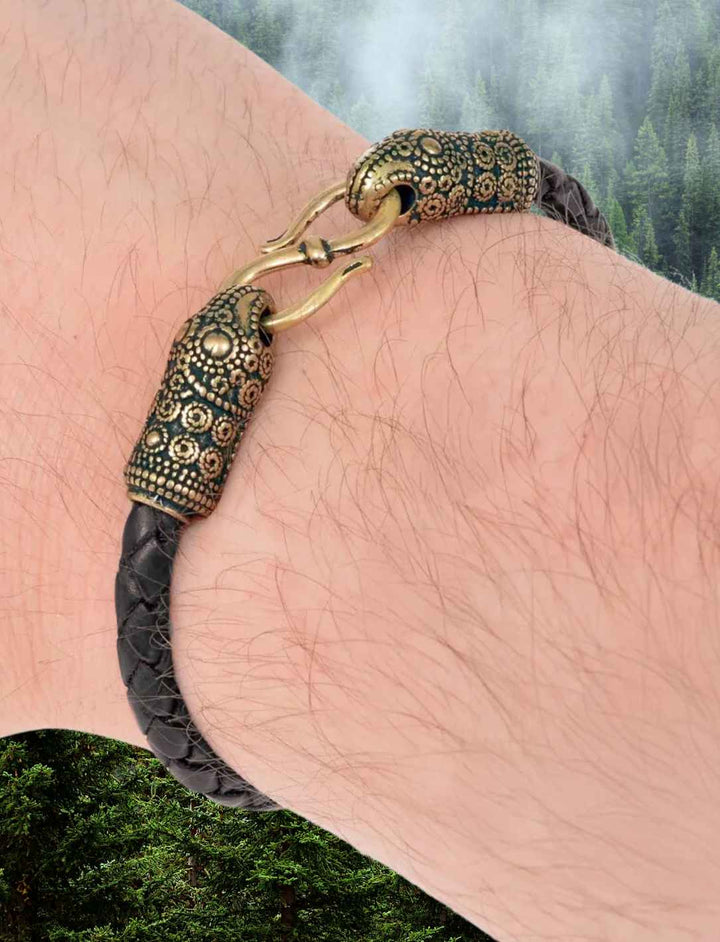 Männerarm, der ein geflochtenes Lederarmband mit bronzefarbenen Rabenköpfen trägt.