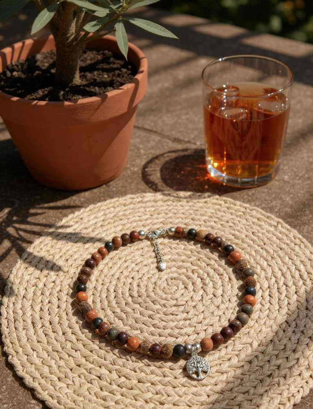 Holzperlen-Choker mit silbernem Lebensbaum-Anhänger, arrangiert auf geflochtener Unterlage neben Glas Tee und Pflanzentopf