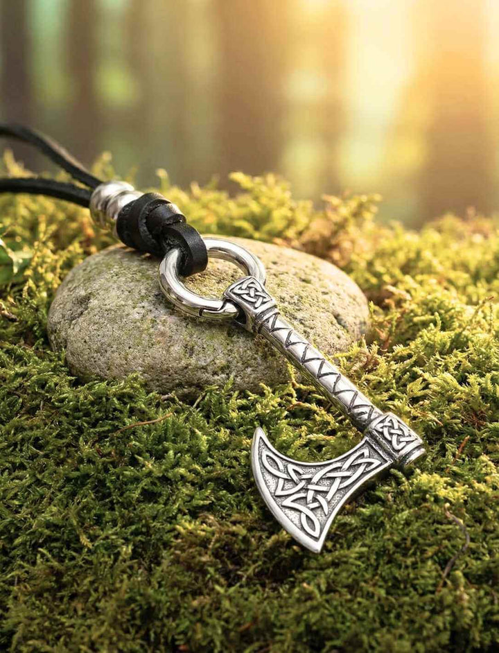 Nordischer Axt Anhänger aus Edelstahl, liegend auf moosigem Waldboden im warmen Licht, mit Lederkette Band des Waldläufers.