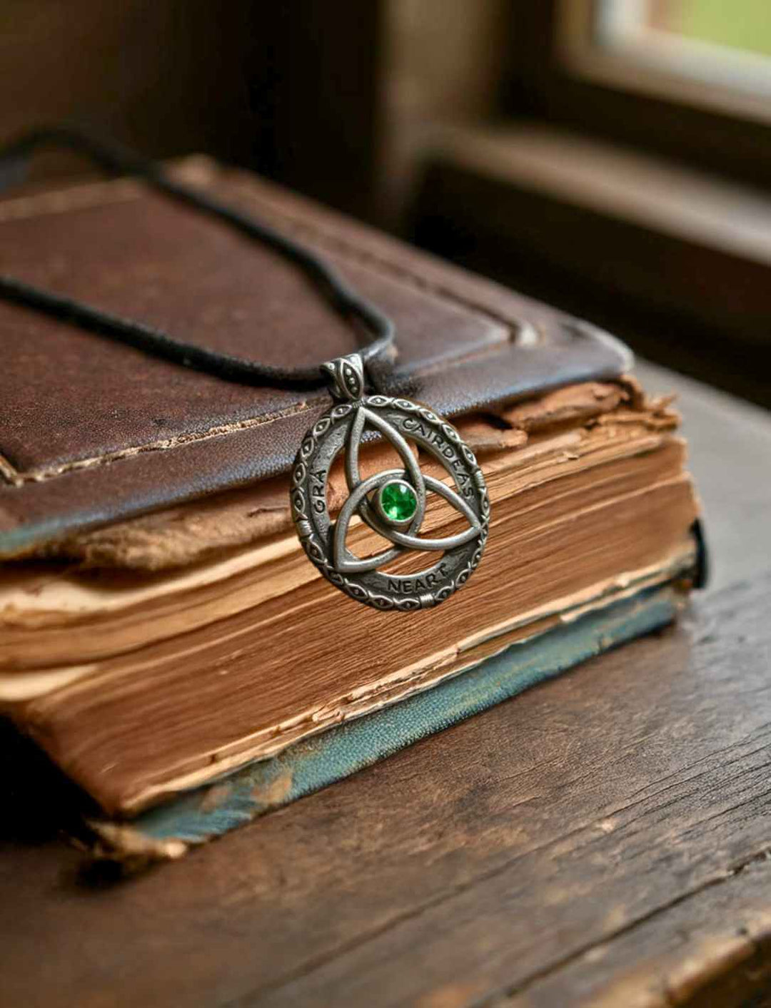 Celtic Bond, Anhänger aus 925 Sterling Silber, präsentiert an einer Lederkette auf einem antiken Buch und Holztisch. In der Mitte ein grüner Zirkona Schmuckstein.